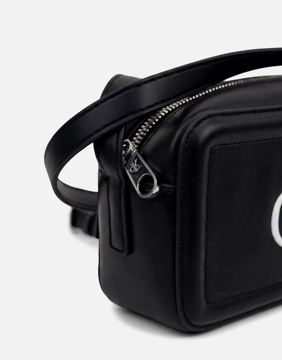 Black Calvin Klein camera bag made of faux leather, featuring a prominent silver zipper with a CK logo pull. Close-up view.