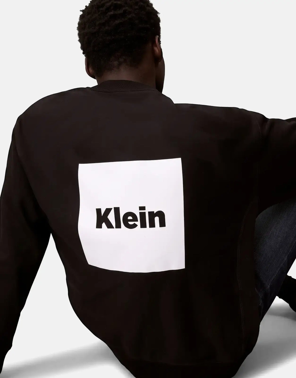 A person from behind wearing a black Calvin Klein Box Graphic Sweatshirt featuring a white square with "Klein" text on the