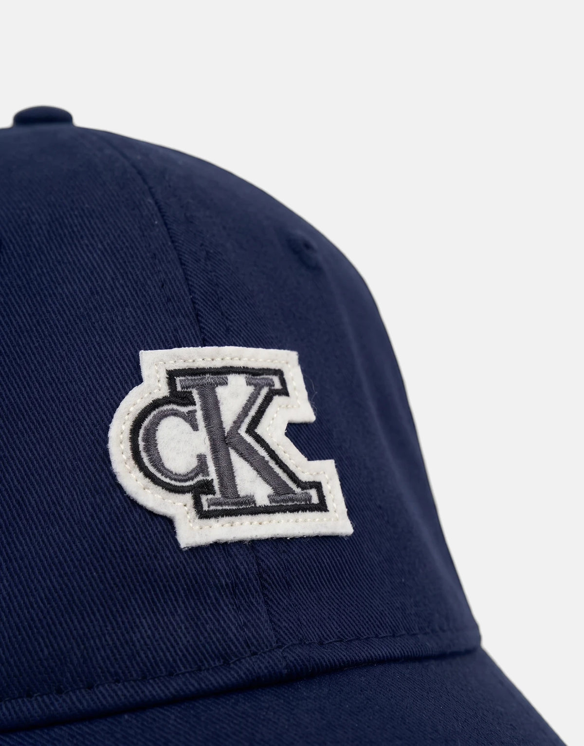 Close-up of a navy blue Calvin Klein cotton twill baseball cap featuring a distinctive white felt 'CK' logo patch on the