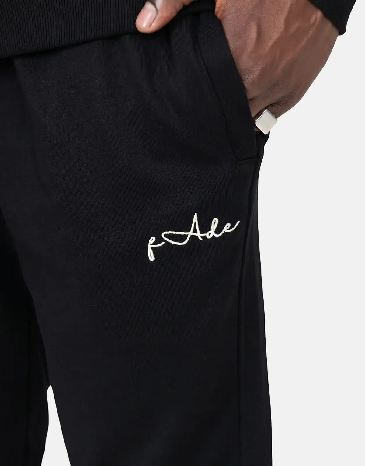 Close-up of black Fade Signature Sweatpants with white embroidered logo, worn by a person with a hand in the pocket,