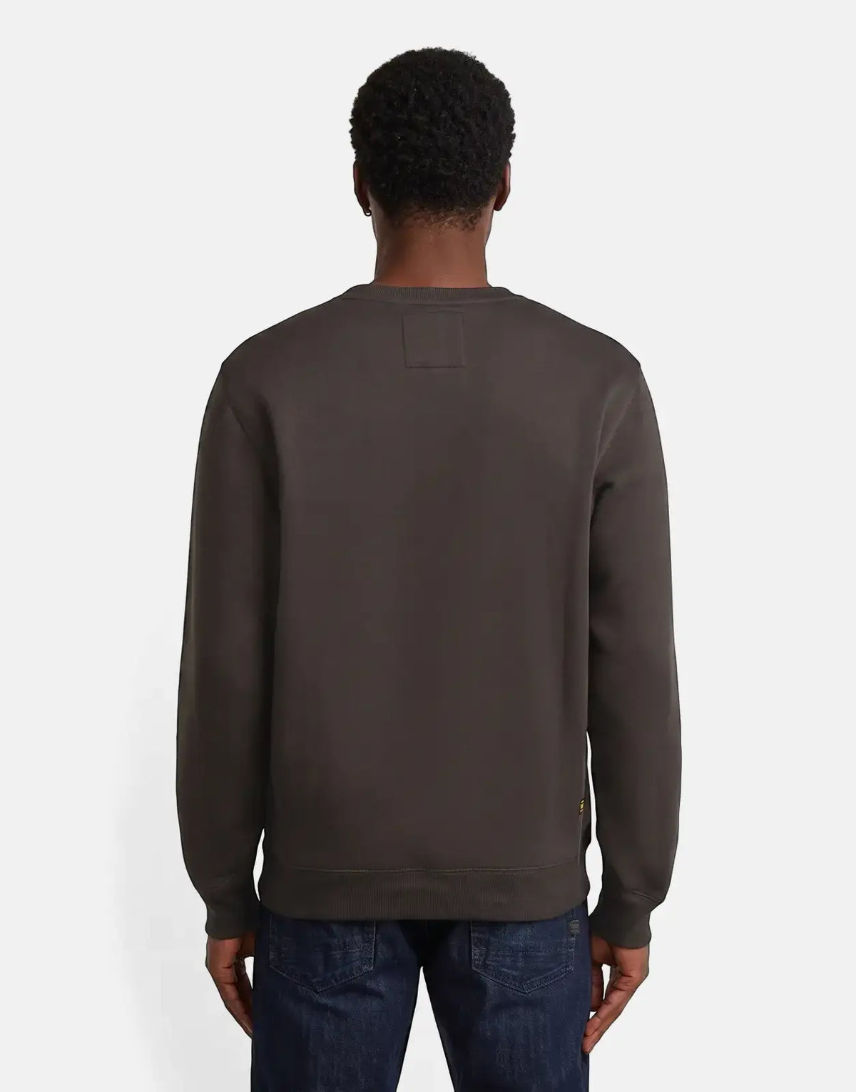 Back view of a male model wearing a dark gray G-Star RAW Premium Core Ganache Sweatshirt and blue jeans, standing on a light