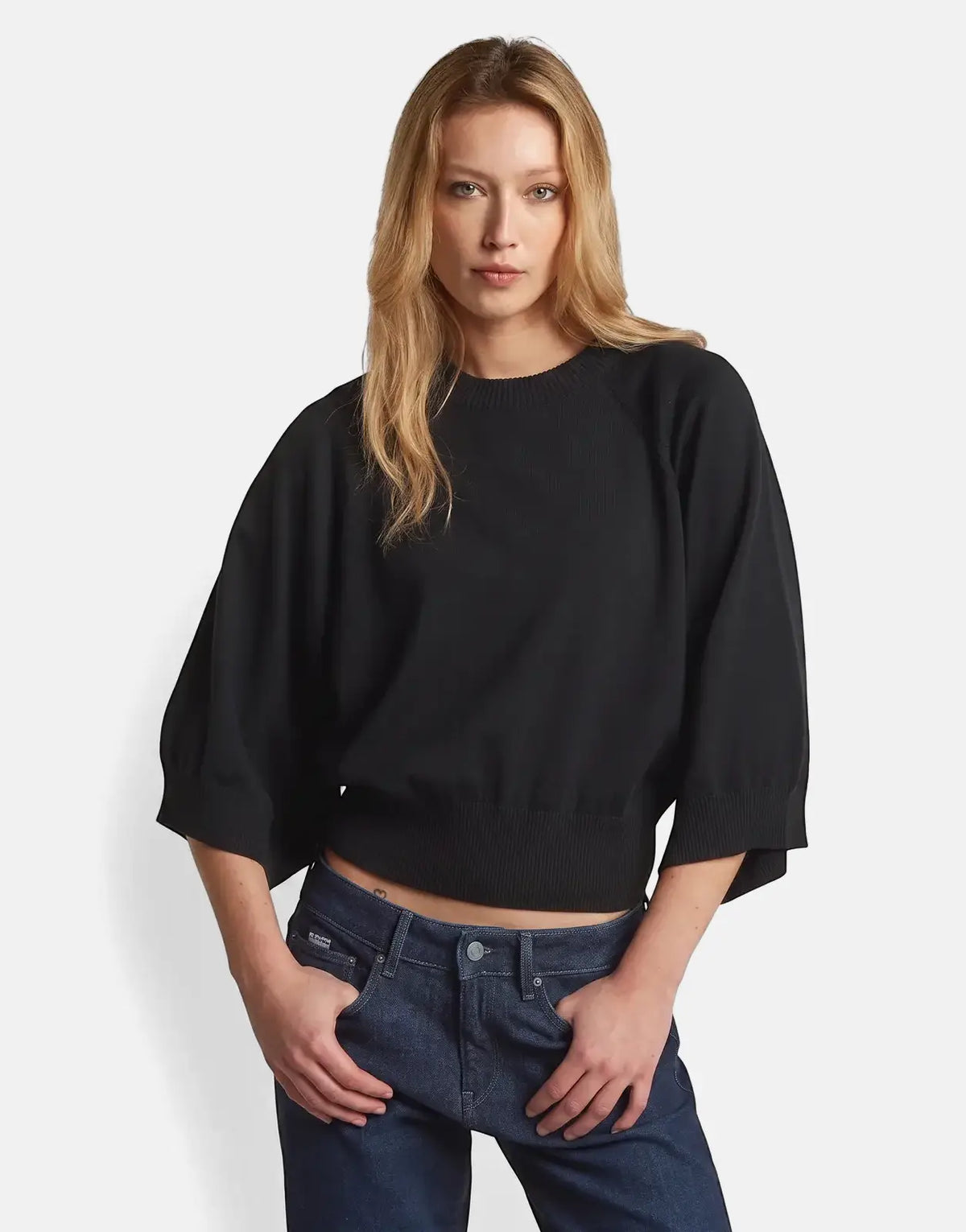 A blonde woman models a G-Star RAW dark black raglan top with wide sleeves and dark wash jeans on a light background.