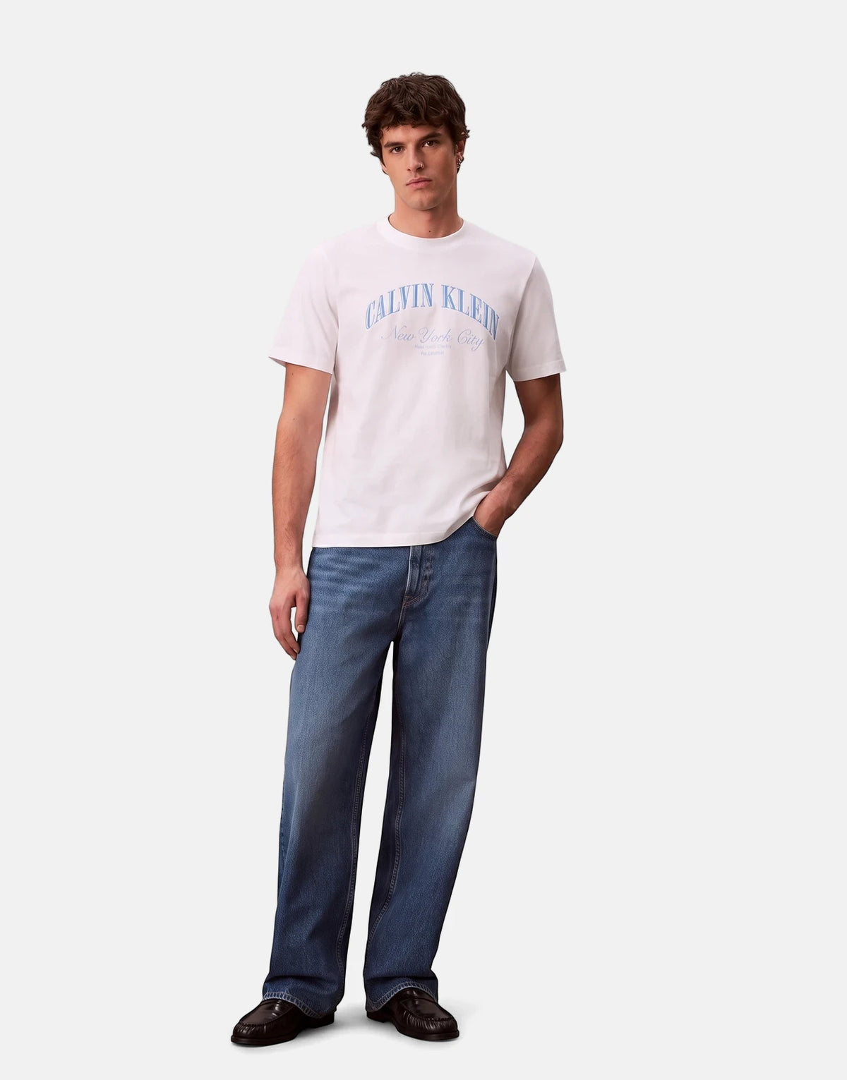 A male model wears a white Calvin Klein graphic t-shirt with blue text and blue relaxed-fit jeans, standing full body.
