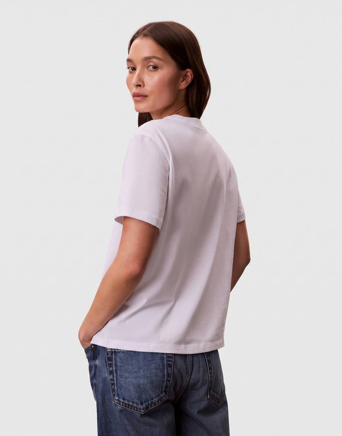 A female model wearing a Calvin Klein Classic Graphic White T-Shirt, shown from the back, paired with blue jeans. The model