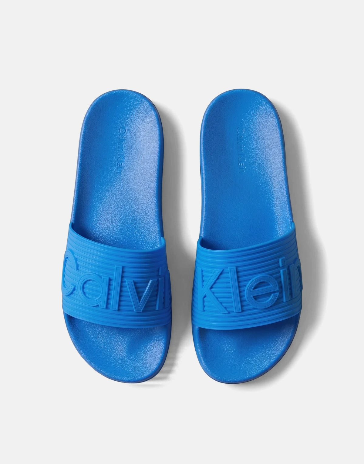 A top-down view of two bright blue Calvin Klein Essential Texture slides with embossed branding, resting on a flat surface.
