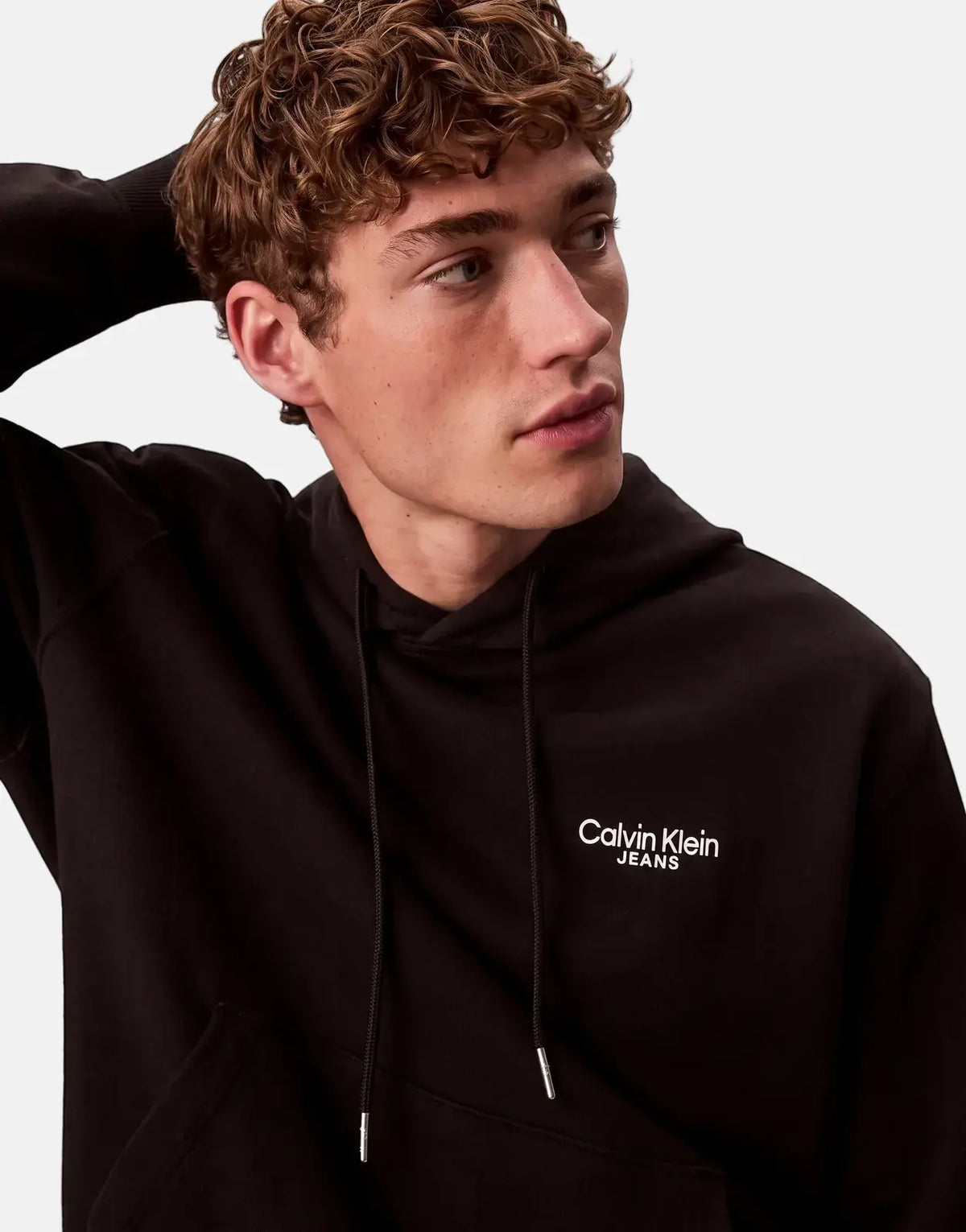 A young man with curly hair models a black Calvin Klein Jeans logo hoodie, posing casually against a plain background.