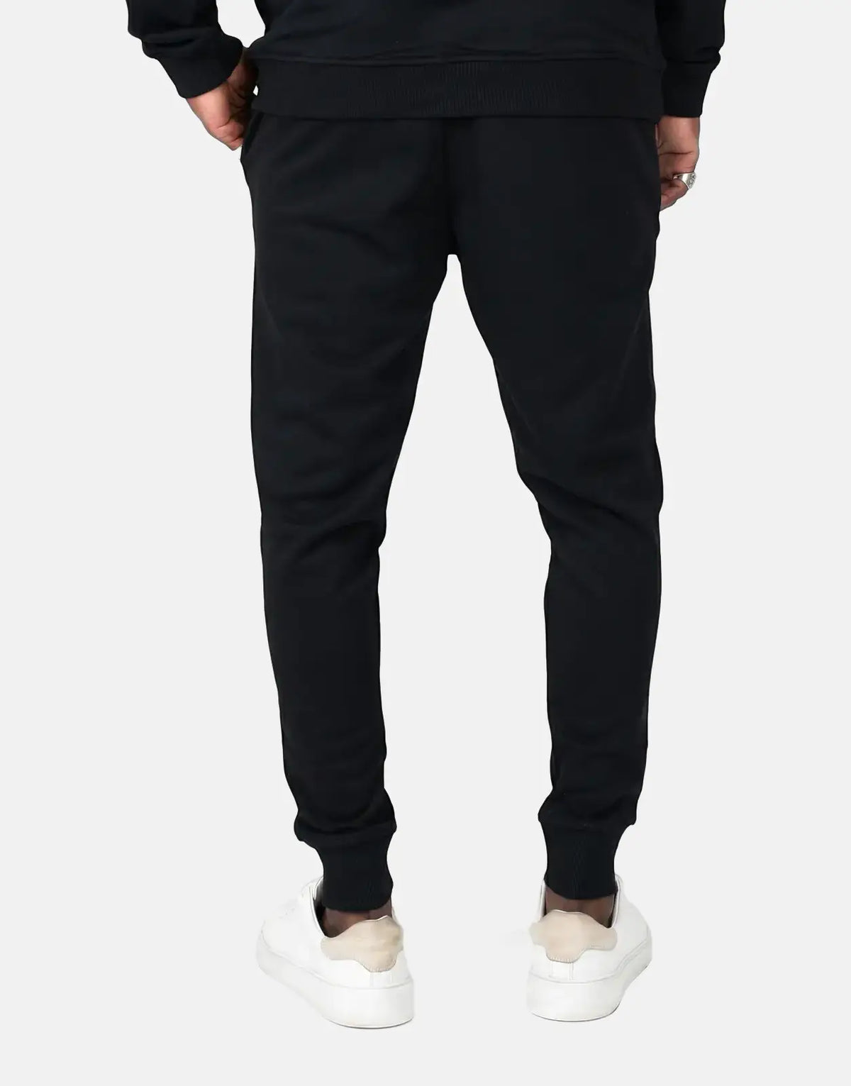 A person from the back wearing black Fade Signature sweatpants with ribbed cuffs and white sneakers on a simple background.