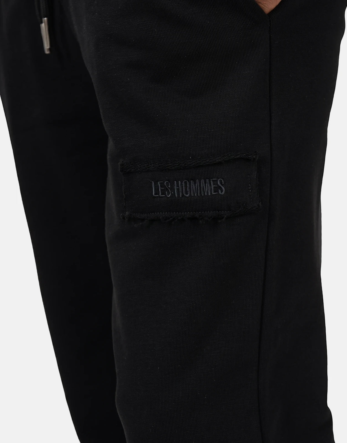 Close-up of Les Hommes black jogger pants featuring a stitched logo label and a silver drawstring tip, side view.